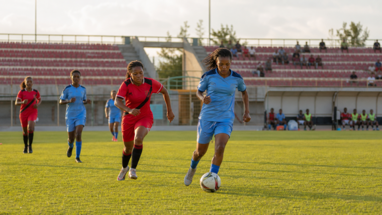 Women play a club football match in a sunlit stadium